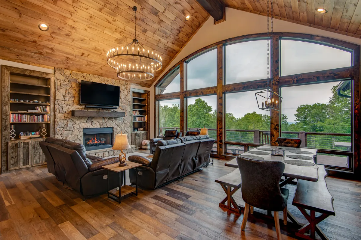 This image captures a spacious, light-filled living and dining area with a stone fireplace, large windows framing scenic mountain views, a chandelier above the dining table, and comfortable leather sofas. The room exudes warmth and invites both relaxation and group gatherings.