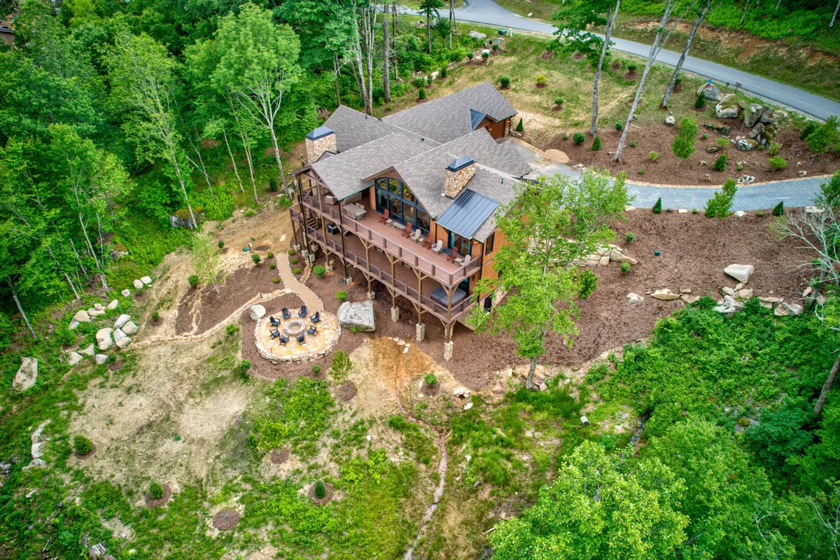 The image showcases the house, an inviting fire pit area surrounded by chairs, and lush trees. The driveway and porch also appear, suggesting multiple gathering spots for both formal team sessions and relaxed, communal evenings under the stars.