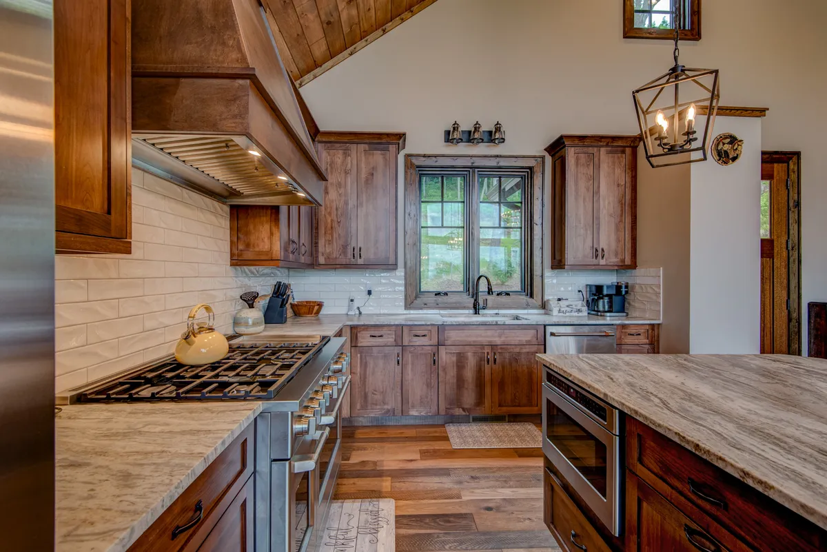 A warm, inviting kitchen with stainless steel appliances, wood cabinetry, a marble countertop, and a large window. The functional and stylish space feels perfect for preparing snacks and drinks while socializing.