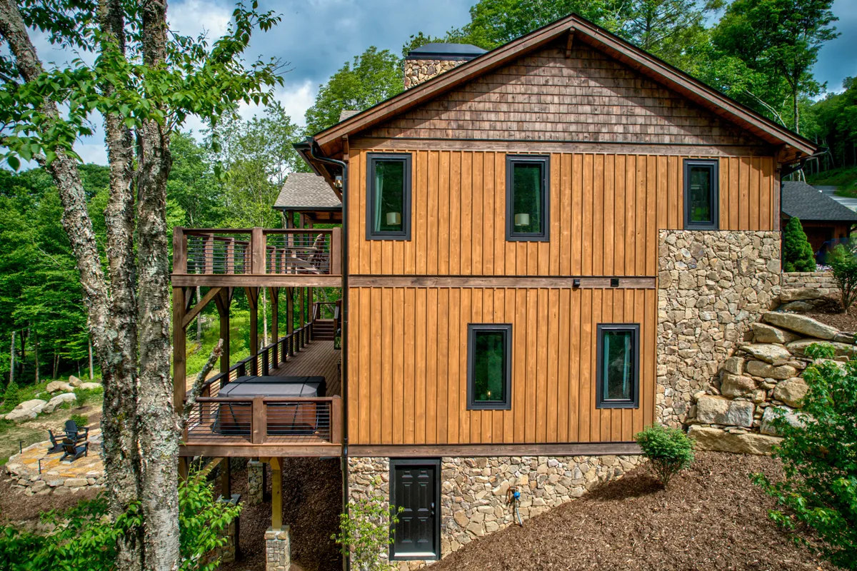 A striking view of the home's wooden facade with a stone base, featuring an expansive deck area surrounded by lush greenery. The scene hints at gatherings on the deck, enjoying nature walks, and relaxing with friends or family. The welcoming atmosphere is highlighted by the soft natural light and open outdoor spaces.