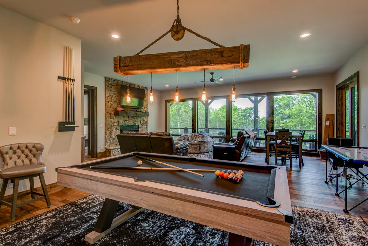 An inviting interior space featuring a pool table, a stone fireplace, comfortable seating, and large windows that bring in beautiful natural light. This game room setting is designed for friendly competition and group entertainment, with an easy flow between indoor and outdoor living.