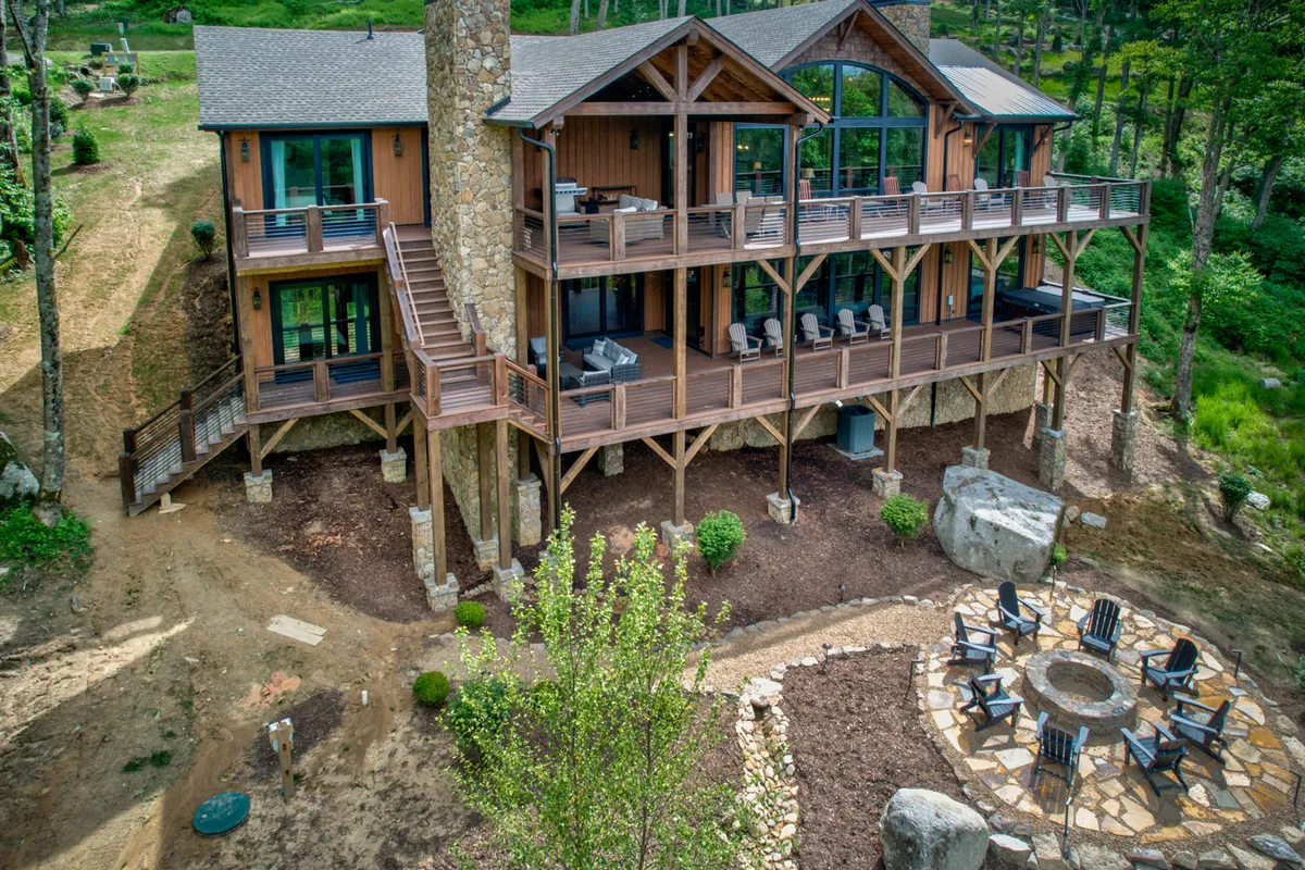 A spacious wooden deck bordered by lush greenery and tall trees, featuring a large stone fire pit surrounded by comfortable lounge chairs. The deck offers ample space for gatherings and relaxation, blending seamlessly with the surrounding natural landscape.