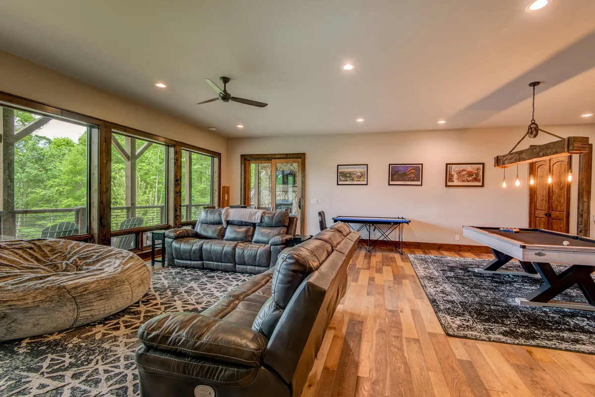 This image captures a casual, welcoming game room featuring a leather sofa, a pool table, a bean bag chair, and large windows that bring in natural light. The view outside emphasizes the seamless connection between indoor entertainment and the outdoors.