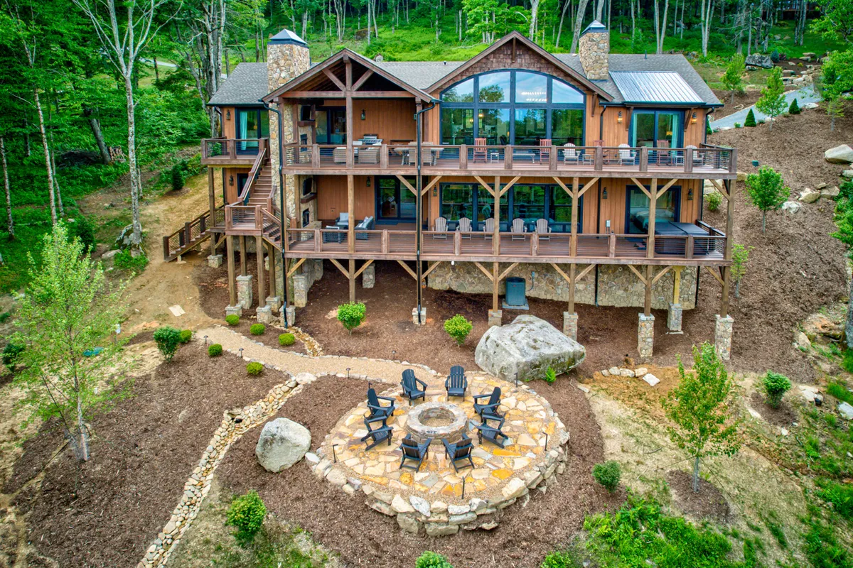 The image showcases the grand wooden cabin set amidst lush mountain greenery, with a spacious circular stone patio featuring a cozy fire pit. Plush outdoor seating invites guests to gather, while the elevated deck promises sweeping sunset views. The scene radiates warmth, togetherness, and a deep connection with nature.