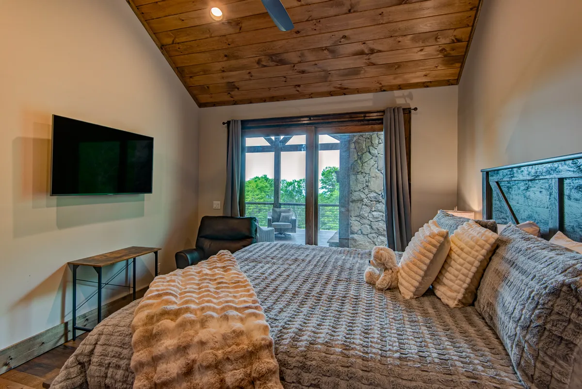 A calm, modern bedroom with a king bed, sliding glass doors that open directly to a private deck, a cozy armchair, and a wall-mounted TV. The scene is filled with soft natural light, suggesting an inviting space for both relaxation and enjoying private outdoor moments.