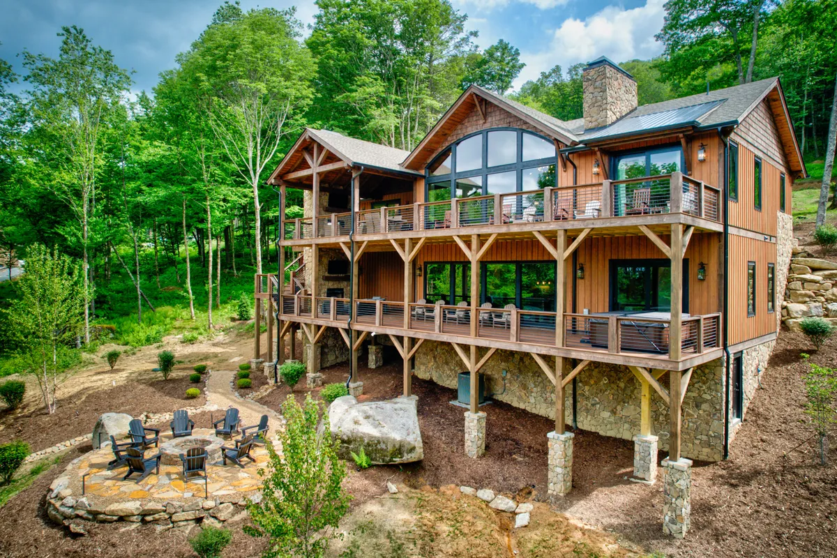 A wide wooden deck and stone patio overlook lush greenery, inviting families to gather for meals, relax by the outdoor fire pit, or explore nearby hiking trails. The expansive windows and outdoor spaces connect the interior comfort with the mountain landscape.