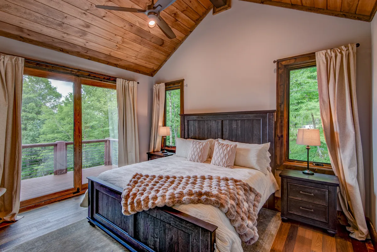 A spacious bedroom featuring a plush king-sized bed dressed in upscale linens, bathed in natural light from large windows that offer relaxing views of surrounding nature. Wooden furniture adds warmth and refinement to the room, creating a serene sanctuary for relaxation.