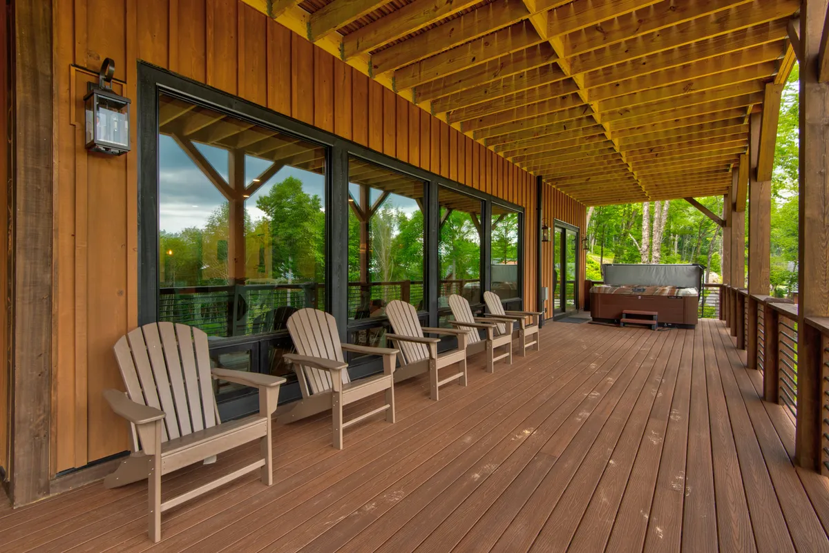 A tranquil deck area features Adirondack chairs, a spacious wooden deck, large windows, and a private hot tub, all oriented towards a beautiful sunset view. The surrounding trees create a peaceful natural enclave ideal for relaxation after a day of adventure.
