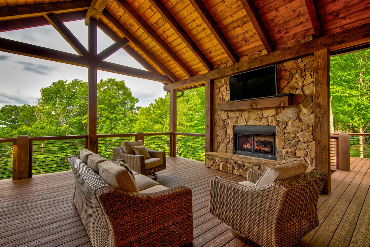 A cozy exterior seating area with a stone fireplace, plush chairs, and expansive windows offering views of the greenery beyond. The space is inviting for both morning coffee and evening relaxation, with direct access to the outdoors.