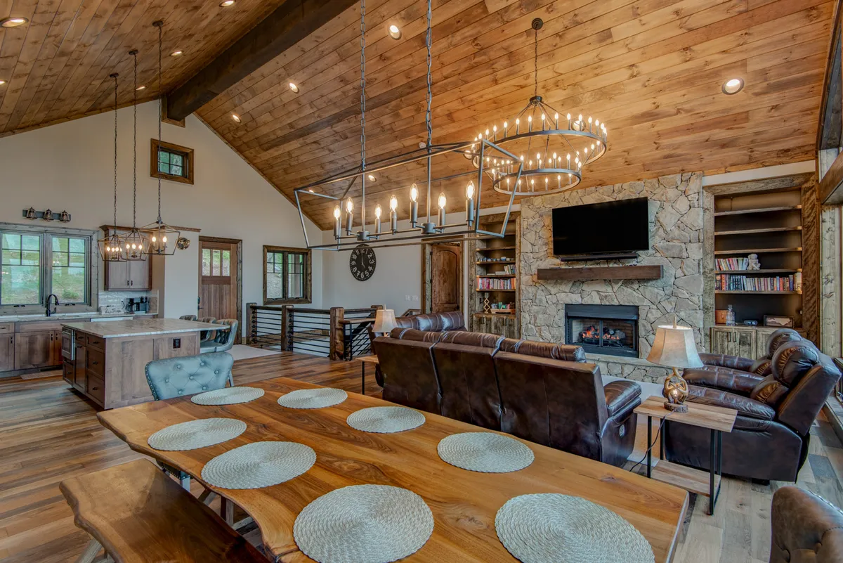 An inviting interior scene showing a chandelier-lit dining table, stone fireplace, and comfortable sofa. The open-plan layout ensures easy movement between dining, lounging, and socializing, all brought together by natural light streaming through the windows.
