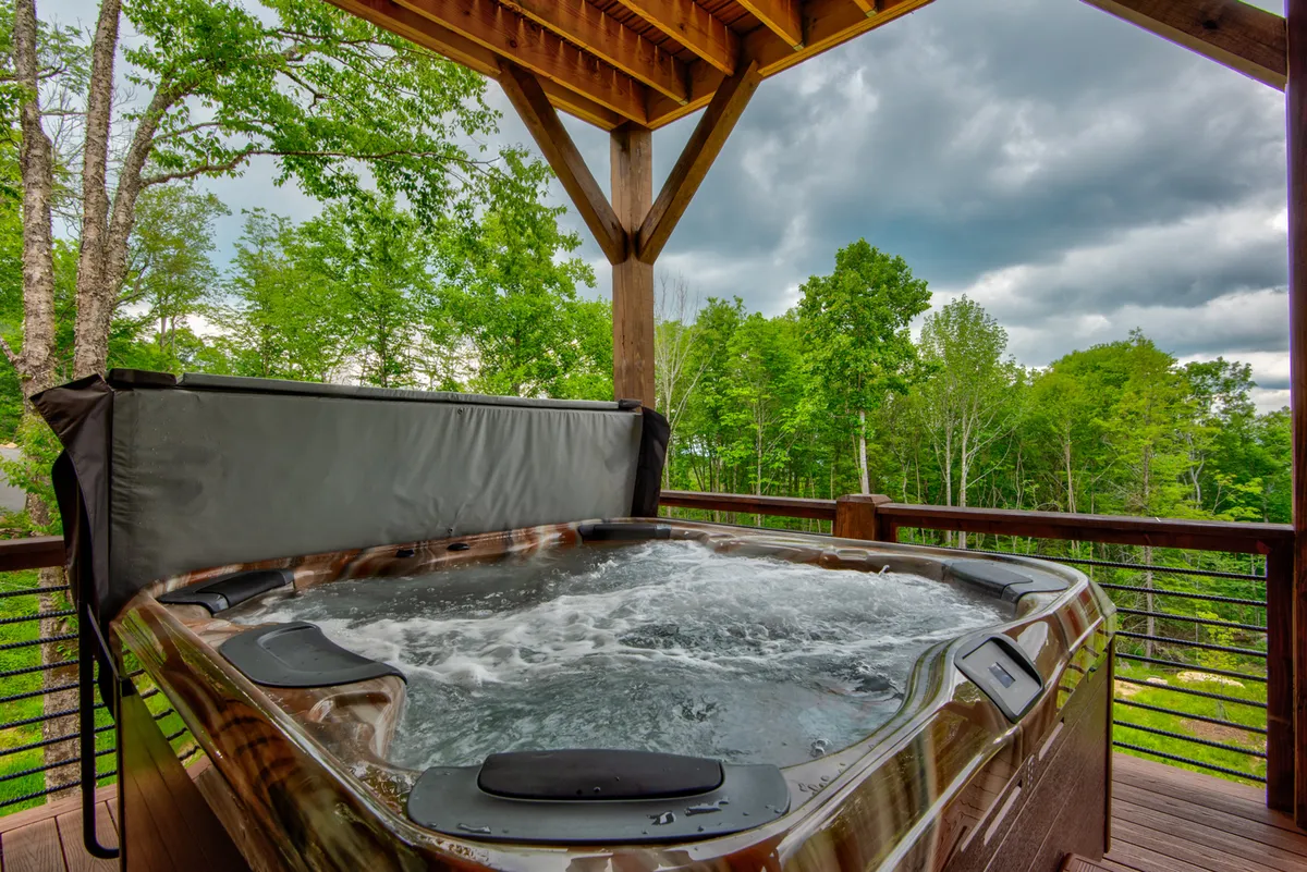 A tranquil scene of a hot tub nestled on the deck, surrounded by towering trees and gentle sunlight filtering through the branches. The inviting hot tub is positioned for privacy and relaxation, with ample room for multiple guests to enjoy mountain air and stargazing together.